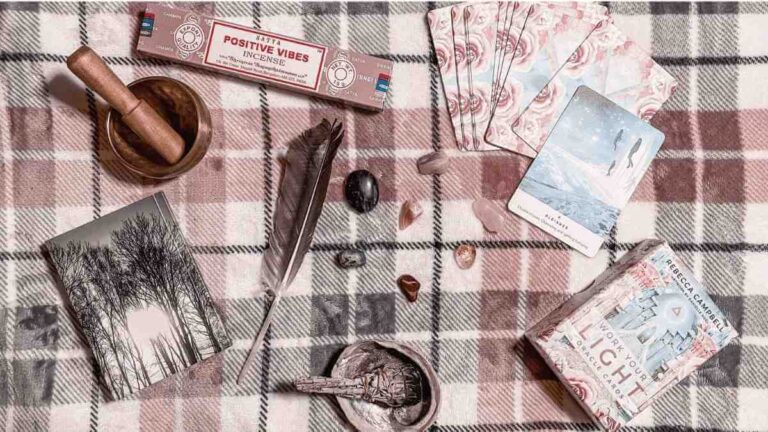 On a pink, white and grey checkered blanket are a selection of healing crystals, a grey notebook with tall trees on it, the Work Your Light oracle cards by Rebecca Campbell, Positive vibes incense sticks in a box, a singing bowl and a sage smudging stick, resting on an Abalone shell and a feather