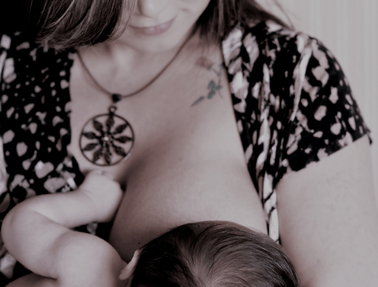 The bottom half of a woman's face, smiling slightly. She is wearing a black and white top and is breastfeeding a newborn baby