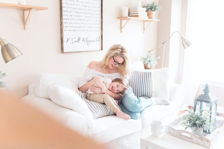 In a living room are shelves with books and a plant. There is a lamp and a white table. On the table is a white mug and an off white tray. On the tray are a plant and a lantern. On the sofa is a woman with blonde hair and glasses. She is wearing jeans and a white top. She is cuddling a toddler and they are both smiling/laughing