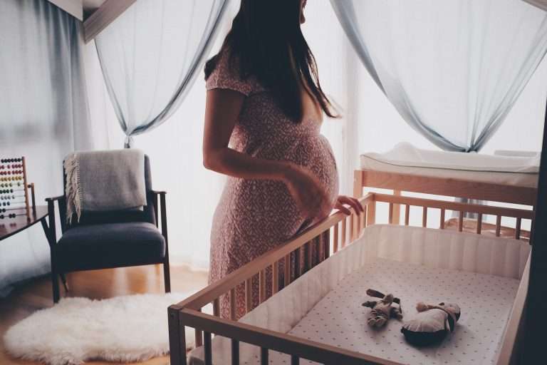 A pregnant woman in a red and white dress is stood over a crib. There are 2 teddies in the crib.