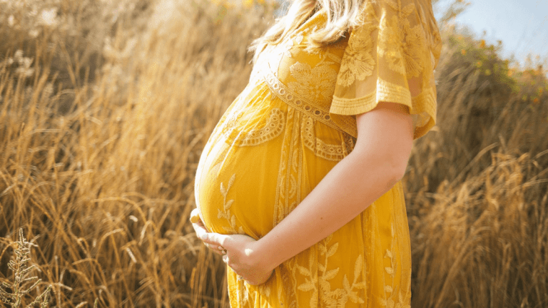 A pregnant woman in a yellow dress is cradling her bump. She outside in the sunshine and is standing in front of tall grass