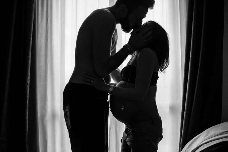 Silhouette of a man and a pregnant woman kissing in front of a window.