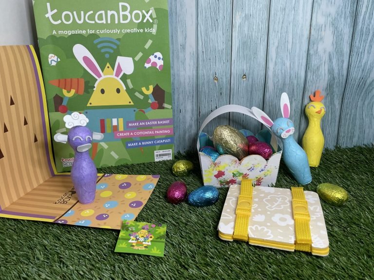 On a grass surface are - a handmade basket with pink, blue and gold egg, a yellow flower press, a toucanBox magazine, 3 small bowling pins in purple (a sheep), blue (a bunny) and yellow (a chick). There is a bowling ally made out of paper and a toucanBox sticker. It is all against a blue fence